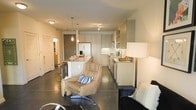 This single-floor space showcases a thoughtfully designed open-concept layout that effortlessly connects the kitchen, dining, and living areas into one cohesive environment. The kitchen is a standout feature, outfitted with flat-front cabinetry in a soft neutral tone that extends floor to ceiling, providing generous storage. A central island with a light stone countertop serves as both a functional prep area and a casual dining spot, complemented by bar-height seating. Pendant lighting with warm amber glass fixtures hangs above the island, adding an inviting glow to the space. Stainless steel appliances, including a French door refrigerator and built-in microwave, lend a polished, contemporary feel. The flooring throughout is polished concrete, offering a sleek, industrial-modern aesthetic that ties the entire space together. The living area features comfortable, modern furnishings, including a plush lounge chair, creating a relaxed yet refined atmosphere. Track lighting and flush-mount ceiling fixtures provide ample illumination throughout. A hallway visible in the background hints at additional private rooms, rounding out this well-appointed and functional single-level layout.