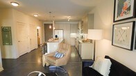 This single-floor space showcases a thoughtfully designed open-concept layout that effortlessly connects the kitchen, dining, and living areas into one cohesive environment. The kitchen is a standout feature, outfitted with flat-front cabinetry in a soft neutral tone that extends floor to ceiling, providing generous storage. A central island with a light stone countertop serves as both a functional prep area and a casual dining spot, complemented by bar-height seating. Pendant lighting with warm amber glass fixtures hangs above the island, adding an inviting glow to the space. Stainless steel appliances, including a French door refrigerator and built-in microwave, lend a polished, contemporary feel. The flooring throughout is polished concrete, offering a sleek, industrial-modern aesthetic that ties the entire space together. The living area features comfortable, modern furnishings, including a plush lounge chair, creating a relaxed yet refined atmosphere. Track lighting and flush-mount ceiling fixtures provide ample illumination throughout. A hallway visible in the background hints at additional private rooms, rounding out this well-appointed and functional single-level layout.