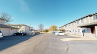 This single-story residential unit is part of a well-maintained multi-building complex set around a spacious shared parking area. The exterior features neutral gray siding with a traditional architectural style, complemented by mature trees and open surroundings visible under wide, clear skies.

Inside, the living room serves as the heart of the unit, finished with wall-to-wall carpeting and neutral-toned walls that create a warm, inviting atmosphere. A large sliding glass door leads to a private patio, allowing natural light to flood the space while offering pleasant outdoor views of open green space. A wall-mounted air conditioning unit provides climate control, and baseboard heating runs along the perimeter.

Adjacent to the living room, the dining area is defined by a glass-top table beneath a modest chandelier, with a decorative mirror and artwork adding character to the clean white walls. The space flows naturally between living and dining zones, making it ideal for everyday use and entertaining.

The kitchen is efficiently laid out in a galley configuration, featuring warm oak cabinetry with ample upper and lower storage, white laminate countertops, and a double-basin sink. White appliances — including a refrigerator, dishwasher, stove, and range hood — are included, and tile flooring provides easy maintenance throughout the cooking space.