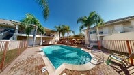 This outdoor pool area is a standout amenity within a gated residential community, offering a well-maintained and inviting space for recreation and relaxation. The centerpiece is a generously sized freeform swimming pool with clear, blue water, accessible via stainless steel entry rails and bordered by a classic brick paver deck that adds warmth and texture to the overall aesthetic.

The pool area is enclosed by a secure metal fence, enhancing both safety and privacy, with a life ring station conveniently mounted along the perimeter. Lounge chairs are positioned poolside, providing comfortable spots to enjoy the abundant natural sunlight that bathes the space throughout the day.

Towering palm trees are strategically placed around the pool, lending a tropical character to the environment and offering dappled shade. A covered pergola structure sits adjacent to the pool deck, providing a shaded seating and gathering area ideal for outdoor entertaining.

The surrounding Mediterranean-influenced residential buildings feature stucco exteriors, tiled rooflines, and private balconies, framing the pool area with architectural cohesion. Covered parking is also visible nearby. Under open blue skies, this outdoor space delivers a resort-like atmosphere that is both functional and visually appealing year-round.