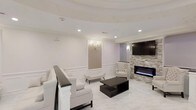 This single-floor space presents a sophisticated and cohesive interior design centered around a neutral white palette. The living area features a tray ceiling with recessed lighting, adding architectural depth and a sense of openness overhead. White wainscoting lines the lower walls, complemented by a decorative framed wall panel adorned with a classic wall sconce, lending a refined, traditional touch to the otherwise contemporary setting.

The seating area is furnished with plush white chairs and a coordinating settee, arranged around a sleek dark espresso coffee table that provides a striking contrast to the light surroundings. A wall-mounted flat-screen television anchors one side of the room, positioned above a stacked stone accent wall housing a modern linear electric fireplace with a blue-flame feature, creating a warm and inviting focal point.

The flooring consists of large-format white marble-style tiles that extend throughout, enhancing the brightness and spaciousness of the area. Together, the blend of classic molding details, contemporary furnishings, and elegant stone accents creates a polished, upscale ambiance throughout the space.