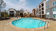 Located at 1701 J N Pease Place in Charlotte, NC, this amenity space is designed to offer a resort-style outdoor experience within a modern residential community. The exterior architecture blends stone, wood-panel siding, and brick accents across multi-story buildings that frame a well-appointed communal area, creating an enclosed yet open atmosphere.

At the heart of the space is a generously sized swimming pool with clear, blue water, surrounded by a spacious stone-tiled deck furnished with lounge chairs and shaded umbrellas. The pool area is complemented by lush landscaping, including mature palm trees and manicured garden beds with low shrubs and ground cover, reinforcing the resort-like ambiance.

A second courtyard area features additional palm trees, paved walkways with decorative railings, built-in seating nooks, and bistro-style tables, offering multiple relaxation zones. The landscaping throughout is thoughtfully designed, incorporating mulched garden beds and greenery that soften the hardscape surroundings.

A covered outdoor lounge pavilion serves as a standout feature, boasting a coffered ceiling with ceiling fans, a wall-mounted television, built-in bench seating, and open slatted wooden screens that allow natural light and outdoor views to filter through. Stone half-walls anchor the structure, and a dining table provides additional gathering space, making it ideal for social events or casual relaxation.