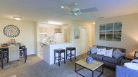 This single-story residence offers an open-concept floor plan that seamlessly connects the kitchen and living areas into one cohesive space. The kitchen is outfitted with light wood cabinetry, white appliances including a refrigerator and range, and a peninsula counter with bar seating for two, making it a practical and social hub. A wine rack detail above the upper cabinets adds a subtle decorative touch. The neutral-toned walls and light-colored cabinetry contribute to a bright, airy feel throughout.

The living area flows naturally from the kitchen, featuring plush carpeted flooring that adds warmth underfoot. A ceiling fan with a light fixture is centrally positioned, providing both air circulation and overhead lighting. The space accommodates comfortable seating with a sofa accented by decorative pillows, complemented by a simple wood-and-metal coffee table. Wall art and a decorative vase introduce coordinated pops of blue. A doorway visible at the rear of the space suggests additional rooms beyond, while the overall layout offers a functional, well-proportioned living environment.