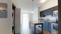 This single-floor space offers a sleek, modern design with an open-concept layout that seamlessly connects the kitchen and living areas. The kitchen is outfitted with dark navy cabinetry, granite countertops, and a classic white subway tile backsplash, creating a striking contrast that anchors the space. A freestanding kitchen island with a matching granite top provides additional prep space and storage, while a stylish pendant light hangs overhead, adding a refined focal point. Wood-look tile flooring runs throughout the unit, offering a warm yet low-maintenance aesthetic. The open corridor leads naturally toward the living area, which is bathed in natural light streaming through a sliding glass door that opens to a private balcony. A ceiling fan is positioned in the living area to ensure comfort year-round. In-unit laundry is conveniently tucked near the entryway, adding a practical touch to the thoughtfully designed layout. The clean white walls and bright, airy atmosphere give the space a fresh, welcoming feel throughout.