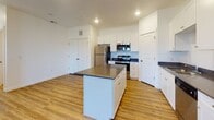 This single-floor unit offers a clean, contemporary layout with thoughtful design throughout. The heart of the home is a well-equipped kitchen featuring crisp white shaker-style cabinetry extending to the ceiling, dark countertops, and a full suite of stainless steel appliances including a refrigerator, range, and over-the-range microwave. A central island with matching dark countertop provides additional prep space and a natural gathering point. Warm-toned hardwood-style flooring runs throughout the main living and kitchen areas, contributing to a bright, cohesive aesthetic. The bedrooms are finished with plush carpeting and are filled with natural light through large windows fitted with blinds, offering outdoor views of the surrounding landscape. A spacious walk-in closet with double hanging rods provides generous storage in the primary bedroom. The bathroom is well-appointed with white cabinetry, a dark countertop vanity with chrome fixtures, a large mirror with overhead lighting, and a full-size soaking tub with shower. Clean white walls and recessed lighting throughout the unit create a fresh, move-in-ready atmosphere that is both functional and inviting.
