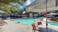 Located at 2700 Central Drive in Bedford, Texas, this outdoor pool area offers a well-appointed communal retreat designed for relaxation and recreation. The centerpiece of the space is a generously sized swimming pool with clear, inviting water, surrounded by a wide pool deck that accommodates multiple lounge chairs and shaded umbrella tables, ideal for sunbathing or casual gatherings.

A charming white gazebo anchors one side of the pool area, providing a shaded architectural focal point that adds a classic, welcoming character to the outdoor environment. Mature trees are thoughtfully distributed throughout the space, offering natural shade and a sense of established greenery that enhances the overall ambiance.

The surrounding landscape is carefully maintained, featuring layered stone retaining walls constructed from natural-toned stacked masonry that add texture and visual interest to the grounds. Ornamental grasses and plantings are integrated throughout, lending a polished, cared-for appearance. Concrete stairways with metal railings provide structured access between the tiered landscaping levels.

The perimeter is secured by a black wrought iron fence, reinforcing safety while maintaining an open, airy feel. The classic brick architecture of the adjacent buildings complements the outdoor design, creating a cohesive and comfortable environment well-suited for community use.