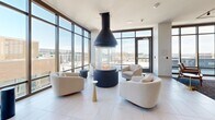 Located in the heart of downtown Madison at 109 E Wilson St, this single-floor combined kitchen and living room space offers a refined, contemporary design with exceptional urban views. Floor-to-ceiling windows wrap the perimeter, flooding the interior with natural light and framing striking panoramic views of the Wisconsin State Capitol and the surrounding cityscape, with direct access to an outdoor terrace.

The living area is anchored by a stunning cylindrical suspended fireplace with a sleek black hood, serving as a dramatic focal point at the center of the room. Comfortable curved seating in neutral tones is arranged around it, creating an inviting and stylish lounge atmosphere. Light-toned large-format tile flooring runs throughout, contributing to the airy, open feel of the space.

The kitchen is equally impressive, featuring an expansive stone-topped island with waterfall edges and warm veining, lined with wooden bar stools beneath a row of striking amber glass pendant lights. Dark-toned open shelving and cabinetry provide generous storage, while premium stainless steel appliances complete the professional-grade setup. A built-in microwave and additional cabinetry in a soft gray tone extend the workspace along the kitchen corridor, connecting seamlessly to the living and dining areas. The overall ambiance is modern, luminous, and meticulously designed.