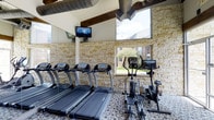 This fitness center offers a thoughtfully designed workout environment that blends functional amenities with distinctive architectural character. The space features striking stacked stone accent walls that lend a rugged, natural texture, complemented by exposed wooden ceiling beams and industrial-style ductwork that give the room an open, loft-like feel. Large windows along the stone wall invite natural light and provide views of the surrounding outdoor greenery, creating an energizing atmosphere for exercise. The flooring is finished with a patterned, rubberized surface well-suited for high-traffic fitness use. The cardio area is outfitted with multiple treadmills, an elliptical machine, and a stationary bike, all arranged to face a wall-mounted flat-screen television for entertainment during workouts. Full-length mirrors are integrated into one wall, enhancing the sense of space and allowing users to monitor their form. The combination of natural materials, ample daylighting, and modern fitness equipment makes this facility a comfortable and motivating space for residents to maintain an active lifestyle.