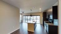 This single-floor space showcases a contemporary open-concept layout that seamlessly connects the kitchen and living area. The dark hardwood flooring runs throughout, providing a rich contrast against the neutral beige walls and white ceilings fitted with recessed lighting.

The kitchen is a functional centerpiece, highlighted by a central island with a built-in sink, light-colored countertop, and stylish pendant lighting overhead. Dark cabinetry lines the perimeter, complementing the stainless steel appliances, including a refrigerator, microwave, and range. The kitchen's design prioritizes both aesthetics and practicality.

The adjacent living area benefits from an abundance of natural light, courtesy of large sliding glass doors that open to a private balcony with exterior views of the surrounding neighborhood. A ceiling fan is installed overhead, enhancing comfort and airflow. The open sightlines between the kitchen and living space create a cohesive, airy atmosphere ideal for both everyday living and entertaining.