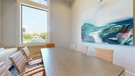 This single-room conference space offers a polished and professional environment designed for focused meetings and collaboration. The room is accessed through a pair of rich, dark wood-framed glass doors that lend both privacy and transparency, allowing natural light to filter through from the interior while maintaining a sense of openness.

At the heart of the room sits a sleek, rectangular conference table surrounded by contemporary upholstered chairs in warm tan and blush tones, providing comfortable seating for small groups. A large, bright window along one wall floods the space with abundant natural light and offers views of the surrounding outdoor greenery, creating an airy and inviting atmosphere.

The neutral, light-toned walls serve as a refined backdrop for a prominent abstract landscape painting rendered in soft blues, greens, and purples — adding a distinctive artistic focal point to the room. Built-in shelving visible through the glass doors in an adjacent area features neatly arranged books and decorative accents, contributing to the overall curated aesthetic.

The flooring transitions between a textured area rug and light hardwood-style surfaces, adding warmth and visual interest underfoot. The space is thoughtfully designed to balance comfort, functionality, and style, making it well-suited for intimate professional gatherings, presentations, or focused work sessions.