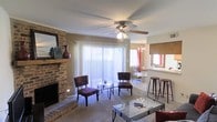 This single-floor residence offers a welcoming and functional layout centered around a comfortable living area. The living room is a standout space, anchored by a full brick fireplace with a classic mantel, providing both a decorative focal point and a cozy gathering spot. A ceiling fan with an integrated light fixture is mounted overhead, promoting airflow and adding to the room's practical charm. Large sliding glass doors dressed with vertical blinds allow abundant natural light to flood the interior while offering access to an outdoor area. The carpeted flooring adds warmth underfoot throughout the living space. Seating is arranged around a glass-topped coffee table, creating an inviting conversation area. Adjacent to the living room, a dining area is visible, illuminated by an elegant chandelier and featuring bar-style seating with tall stools along a counter or pass-through, suggesting a convenient connection to the kitchen. A secondary window with red accent curtains adds a pop of color to the dining space. Overall, the home presents a traditional, well-appointed interior that balances comfort and everyday functionality.