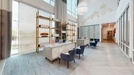 Located at 375 54th Ave N in Saint Petersburg, FL, this sophisticated single-floor amenity space spans a generous footprint and delivers a polished, resort-caliber experience. The grand lobby serves as the centerpiece, boasting soaring double-height ceilings, floor-to-ceiling glass walls, and an abundance of natural light. A sleek reception desk clad in veined stone is flanked by gold-accented open shelving, navy blue accent chairs, and woven pendant light fixtures, creating an inviting yet professional first impression. Throughout the space, light-toned hardwood flooring in a herringbone pattern complements a neutral, coastal-inspired palette accented with deep blues, warm golds, and crisp whites. Multiple lounge areas offer varied seating arrangements, including a sitting area anchored by a dramatic full-height marble feature wall with an integrated linear fireplace and wall-mounted display screen. A grand piano adds a cultural focal point to the open social lounge. A dedicated conference room features large windows overlooking tropical landscaping, a sleek meeting table, and a wall-mounted screen alongside a decorative moss installation. A café-style bar area with dark cabinetry, a white countertop island, gold pendant lighting, and patterned wallcovering provides a functional refreshment hub. Panoramic views of the pool and waterway are visible throughout, enhancing the bright, open, and luxurious ambiance of this exceptional amenity space.