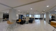 This single-floor space presents a contemporary open-plan lounge or lobby area designed for comfort and functionality. The flooring features large-format marble-look tiles that lend a polished, upscale aesthetic throughout. A striking dropped ceiling detail with a curved edge adds architectural interest overhead, while an abundance of recessed lighting ensures the space is evenly and warmly illuminated.

A standout feature is the pair of sleek, modern linear fireplaces positioned side by side along a central wall, serving as a focal point for the room. Multiple seating arrangements surround this area, including dark upholstered armchairs and sofas grouped around low coffee tables, offering versatile options for gathering or relaxing. A wall-mounted flat-screen television is positioned above the fireplaces for added functionality.

Natural light filters in through several windows along the perimeter, offering views of surrounding greenery. Toward the rear of the space, additional seating nooks provide more intimate settings. The overall ambiance is clean, modern, and inviting, balancing open communal space with defined seating zones suitable for both social interaction and quiet relaxation.
