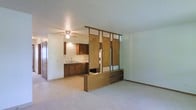 This single-floor space offers a practical and functional layout defined by its notable design features. The living area is finished with wall-to-wall carpet, providing a soft and comfortable underfoot feel, while a textured ceiling adds a classic touch overhead. A ceiling fan with integrated lighting serves as the central fixture, ensuring both airflow and illumination throughout the open space.

A standout architectural element is the decorative wooden room divider that partially separates the living area from the kitchen. This divider features vertical panels and a built-in stepped platform at its base, adding visual interest and a sense of definition between the two spaces without fully enclosing either area.

The kitchen is appointed with warm-toned wood cabinetry along both the upper and lower sections, offering ample storage. A single-basin sink is positioned beneath the cabinetry, and the layout provides functional counter space for everyday use. The kitchen area transitions smoothly into the living space, reinforcing the open-concept feel of the floor plan.

An interior door is visible along the wall, suggesting additional rooms or storage areas beyond the main open space. Overall, the layout balances openness with subtle spatial definition, making it a versatile and livable single-floor environment.