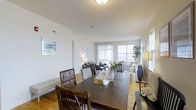 This two-story loft at Summerfield Lofts offers a thoughtfully designed living space that combines comfort with an airy, modern aesthetic. The main level showcases an open-plan layout where the dining and living areas flow seamlessly into one another, creating a cohesive and inviting environment. Warm hardwood flooring runs throughout, anchoring the space with a natural, welcoming tone.

The dining area features a substantial dark wood table paired with rattan and striped upholstered chairs, providing a stylish yet functional gathering space. Adjacent to the dining area, the living room is generously proportioned, accommodating a full seating arrangement with ease. Large windows line the perimeter, flooding the interior with natural light and offering bright, open views that enhance the sense of spaciousness.

The clean white walls serve as a neutral backdrop, complemented by tasteful artwork and greenery that add warmth and personality. Recessed and ambient lighting supplement the natural light, ensuring the space remains well-lit throughout the day. The overall ambiance is bright, calm, and contemporary, reflecting a balanced blend of functional design and comfortable living across both floors of this Summerfield Lofts residence.