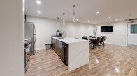 Located at 2911 Bayview in Toronto's M2K neighbourhood, this thoughtfully designed amenity space offers a generously sized, open-concept layout spread across a single floor. Warm-toned hardwood flooring runs throughout, unifying the various functional zones and reflecting the recessed pot lighting that keeps the space bright and welcoming.

The open living area features multiple seating arrangements, including a plush sectional sofa, cozy armchairs, and a circular glass coffee table, all anchored by a sleek wall-mounted electric fireplace with a dramatic flame display. Adjacent to this is a secondary lounge area furnished with rich leather club chairs, ideal for relaxed conversation or quiet gatherings.

The kitchen is a standout feature, showcasing a large island with a striking marble-effect quartz countertop, dark cabinetry, stainless steel appliances, and elegant pendant lighting overhead. A dedicated dining area nearby accommodates a full-sized table with upholstered chairs and includes a wall-mounted television.

A welcoming entry zone with double glass doors and additional seating completes the layout. Neutral walls, consistent flooring, and carefully chosen furnishings give the entire space a cohesive, contemporary ambiance suited for community use or social gatherings.