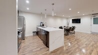 Located at 2911 Bayview in Toronto's M2K neighbourhood, this thoughtfully designed amenity space offers a generously sized, open-concept layout spread across a single floor. Warm-toned hardwood flooring runs throughout, unifying the various functional zones and reflecting the recessed pot lighting that keeps the space bright and welcoming.

The open living area features multiple seating arrangements, including a plush sectional sofa, cozy armchairs, and a circular glass coffee table, all anchored by a sleek wall-mounted electric fireplace with a dramatic flame display. Adjacent to this is a secondary lounge area furnished with rich leather club chairs, ideal for relaxed conversation or quiet gatherings.

The kitchen is a standout feature, showcasing a large island with a striking marble-effect quartz countertop, dark cabinetry, stainless steel appliances, and elegant pendant lighting overhead. A dedicated dining area nearby accommodates a full-sized table with upholstered chairs and includes a wall-mounted television.

A welcoming entry zone with double glass doors and additional seating completes the layout. Neutral walls, consistent flooring, and carefully chosen furnishings give the entire space a cohesive, contemporary ambiance suited for community use or social gatherings.