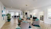 Located at 1 Cedarwood Way in Newport News, Virginia, this single-floor community clubhouse offers a well-appointed and versatile gathering space. The open-concept living and dining area serves as the heart of the space, featuring warm wood-tone flooring, recessed lighting, and an elegant chandelier that anchors the room. The living area is furnished with contemporary lounge seating accented by teal throw pillows, arranged around a central coffee table atop a geometric area rug, creating an inviting and cohesive aesthetic. Decorative columns define the transition between the living and dining zones, where a formal dining table with modern chairs sits beneath a matching chandelier and is bathed in natural light from multiple windows. A kitchen with dark cabinetry and stainless steel appliances is accessible from the main living area, providing functional support for events and gatherings. The fitness center is a standout feature, offering a comprehensive range of equipment including treadmills, cable machines, a cable crossover station, free weights, and an elliptical, all set on durable rubber flooring. Natural light filters in through multiple windows, and wall-mounted mirrors enhance the sense of space. The soft sage-green walls of the exercise room add a calm, motivating atmosphere throughout.