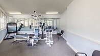 Located at 3540 NW 50th Ave in Lauderdale Lakes, Florida, this fitness center offers a single, well-equipped exercise room designed to support a comprehensive workout experience. The space is characterized by an abundance of natural light streaming through large, multi-pane windows that line the walls and provide pleasant views of the surrounding tree-filled outdoor landscape. Full-length wall mirrors amplify the sense of openness and allow users to monitor their form during exercise. The room features a Paramount multi-station strength training machine with cable pulleys, weight stacks, a leg press, and bench attachments — suitable for a wide range of resistance exercises. Additional equipment includes a treadmill, a free-weight barbell with weight plates, and a dedicated weight bench. The flooring is covered in durable gray carpet, well-suited for a fitness environment, while the textured ceiling is fitted with fluorescent lighting to ensure consistent illumination throughout. The light blue-gray painted walls and clean, minimalist layout contribute to a calm yet functional atmosphere. Seating chairs are available along the perimeter, offering a convenient rest area between sets. Overall, this fitness center provides a practical and well-lit space for both strength training and cardio exercise.