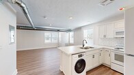 Located at 1542 Michigan St NE in Grand Rapids, Michigan, this single-floor unit has been thoughtfully renovated with a clean, modern aesthetic. The open-concept layout connects the living area to the kitchen, creating a bright and functional space filled with natural light from multiple windows fitted with white blinds. Light wood-tone luxury vinyl plank flooring runs throughout, tying each area together seamlessly.

The kitchen is a standout feature, showcasing white shaker-style cabinetry, a classic white subway tile backsplash, and a peninsula with quartz countertops that doubles as a prep and dining area. A stainless steel sink with a matte black faucet is integrated into the peninsula. The kitchen is equipped with white appliances, including a refrigerator, range, and built-in microwave, along with the added convenience of an in-unit washer tucked neatly beneath the counter.

The living and bedroom areas offer open, flexible spaces with exposed silver ductwork running along the ceiling, adding an industrial character to the otherwise clean interior. Recessed lighting provides warm illumination throughout.

The bathroom continues the cohesive design with white shaker vanity cabinetry, a quartz countertop, matte black hardware and fixtures, a round mirror, and wood-look flooring. The space is bright and well-appointed, reflecting the same attention to detail found throughout the unit.