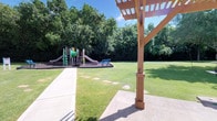 This well-maintained outdoor space offers a thoughtfully designed recreational environment suited for community gathering and play. A sturdy wooden pergola anchors one side of the area, providing shaded relief and a defined gathering point beneath its open-beam structure. From here, a smooth concrete pathway extends across the grounds, complemented by stepping stone accents that guide visitors across the manicured green lawn.

The centerpiece of the space is a colorful children's playground, featuring multiple slides, climbing structures, and play elements set within a protective mulch-covered safety surface. A bench positioned near the playground offers a convenient spot for supervision and rest. The play area is generously sized and designed to accommodate active play for a range of ages.

Mature, leafy trees line the perimeter of the space, creating a natural green boundary that enhances privacy, reduces ambient noise, and provides additional shade throughout the day. The combination of open lawn areas and structured features gives this space a balanced, versatile character. Abundant natural light floods the grounds on clear days, further enhancing the inviting and energetic atmosphere of this outdoor recreational area.