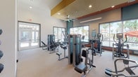 This single-floor fitness center is designed to deliver a complete workout experience in a contemporary, well-appointed setting. The main gym floor is generously proportioned, featuring soaring ceilings with bold teal and terracotta accent panels that add architectural character. Large picture windows line the perimeter, flooding the space with natural light and offering views of palm trees, a pool, and a resort-style outdoor amenity area beyond. A ceiling fan and recessed lighting ensure year-round comfort throughout the workout area.

The cardio zone is equipped with multiple treadmills, ellipticals, and a rowing machine, all positioned to take advantage of the scenic outdoor views. The strength training section includes a full rack of dumbbells, multiple cable and resistance machines, adjustable benches, and wall-mounted mirrors that enhance the sense of space. Mounted flat-screen televisions are positioned throughout for entertainment during workouts.

A dedicated studio room features rich hardwood flooring, a tray ceiling with ambient LED lighting, a wall-mounted television, and textured accent wall panels — ideal for yoga, stretching, or personal training sessions. A utility area provides dual drinking fountains, a countertop station with cabinetry, and a window overlooking the exterior. Rubber-composite flooring throughout the main gym area ensures durability and safety.
