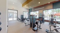 This single-floor fitness center is designed to deliver a complete workout experience in a contemporary, well-appointed setting. The main gym floor is generously proportioned, featuring soaring ceilings with bold teal and terracotta accent panels that add architectural character. Large picture windows line the perimeter, flooding the space with natural light and offering views of palm trees, a pool, and a resort-style outdoor amenity area beyond. A ceiling fan and recessed lighting ensure year-round comfort throughout the workout area.

The cardio zone is equipped with multiple treadmills, ellipticals, and a rowing machine, all positioned to take advantage of the scenic outdoor views. The strength training section includes a full rack of dumbbells, multiple cable and resistance machines, adjustable benches, and wall-mounted mirrors that enhance the sense of space. Mounted flat-screen televisions are positioned throughout for entertainment during workouts.

A dedicated studio room features rich hardwood flooring, a tray ceiling with ambient LED lighting, a wall-mounted television, and textured accent wall panels — ideal for yoga, stretching, or personal training sessions. A utility area provides dual drinking fountains, a countertop station with cabinetry, and a window overlooking the exterior. Rubber-composite flooring throughout the main gym area ensures durability and safety.