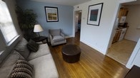 This single-floor residence offers a comfortable and well-organized living environment with a warm, inviting ambiance throughout. The living room serves as the heart of the space, showcasing rich hardwood flooring that adds a timeless character to the interior. Soft blue-gray walls create a calm, relaxing atmosphere, complemented by white trim and baseboards that provide a clean, crisp contrast. The room is furnished with a plush sofa, an upholstered armchair, and a round leather ottoman, offering ample seating and a cozy setting for everyday living. Framed artwork adorns the walls, adding thoughtful decorative touches that enhance the overall aesthetic. A side table with a table lamp provides warm, ambient lighting, while a lush indoor plant in the corner introduces a natural element to the space. A doorway visible from the living area leads to additional rooms, including what appears to be a bedroom, suggesting a practical and connected floor plan. The layout flows naturally, making efficient use of the available space. Overall, the residence presents a neat, move-in-ready interior with classic finishes and a welcoming character throughout.