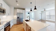 This well-designed 2-bedroom, 2-bathroom residence offers a thoughtful, modern layout across a single floor. The open-concept kitchen and living area serves as the heart of the home, featuring white shaker cabinetry extending to the ceiling, a classic subway tile backsplash, and a full suite of stainless steel appliances including a French door refrigerator, range, and built-in microwave. A central island with a white quartz countertop, undermount sink with a matte black faucet, and stylish dual pendant lights make this kitchen both functional and visually striking. Light wood-tone flooring flows throughout the main living areas, complementing the bright, white walls and creating a clean, contemporary aesthetic. The living area benefits from multiple windows that invite generous natural light and outdoor views. Both bedrooms are carpeted and include ceiling fans and walk-in closets equipped with shelving and hanging rods. The primary bathroom features a dual-sink vanity with white countertops, matte black fixtures, a large wall mirror with contemporary lighting, and a glass-enclosed shower. A dedicated hallway connects the rooms efficiently. The overall design is cohesive and modern, combining neutral tones, quality finishes, and practical storage throughout.