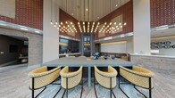 This single-floor communal space combines industrial and contemporary design elements to create a welcoming, multi-functional lounge environment. The defining architectural feature is the dramatically tall ceiling, accentuated by an expansive installation of cascading Edison-style pendant bulbs that draw the eye upward and fill the space with warm ambient light. Exposed brick walls in rich red tones contrast with smooth grey columns and white upper walls, lending the interior a refined industrial character.

The flooring throughout is a natural stone tile in warm earth tones, contributing to an inviting, grounded atmosphere. At the center of the space sits a substantial conference-style table in slate blue-grey, surrounded by upholstered accent chairs with bold geometric yellow patterning, providing an ideal setting for meetings or collaborative gatherings.

Along one wall, a wet bar or kitchenette with dark cabinetry and glossy tile backsplash serves as a functional amenity hub. A mounted display screen nearby adds a media-ready element to the space. Toward the perimeter, varied lounge seating—including leather benches, plaid accent chairs, and cushioned armchairs—creates intimate conversation areas. Large floor-to-ceiling windows and glass entry doors flood the space with natural light while offering clear views of the outdoor streetscape, enhancing the connection between interior and exterior.