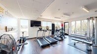 Located at 6711 NE 182nd St in Kenmore, WA, this fitness center offers a well-appointed exercise space designed to support a comprehensive workout experience. The single-floor layout is thoughtfully organized to maximize functionality while maintaining an open, airy feel.

Large windows along multiple walls flood the room with abundant natural light and provide views of the surrounding greenery and neighboring building exteriors, creating an energizing atmosphere. The space features durable hardwood-style flooring throughout, complemented by protective rubber mats positioned beneath the equipment.

The cardio section is equipped with multiple treadmills and elliptical trainers from TRUE Fitness, as well as a rowing machine and stationary bike. A multi-station cable strength machine anchors the strength training area, accompanied by a weight bench, medicine balls, and a wall-mounted rack of dumbbells. Stability and exercise balls add versatility for functional training.

A striking motivational word-art mural decorates one wall, displaying fitness-themed phrases such as "aim higher," "commitment," and "dedication," adding an inspiring visual element to the space. Recessed and linear LED lighting in the tray-style ceiling ensures consistent illumination throughout. A wall-mounted flat-screen television provides entertainment during workouts. The overall design is clean, modern, and purpose-built for an effective fitness experience.