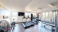 Located at 6711 NE 182nd St in Kenmore, WA, this fitness center offers a well-appointed exercise space designed to support a comprehensive workout experience. The single-floor layout is thoughtfully organized to maximize functionality while maintaining an open, airy feel.

Large windows along multiple walls flood the room with abundant natural light and provide views of the surrounding greenery and neighboring building exteriors, creating an energizing atmosphere. The space features durable hardwood-style flooring throughout, complemented by protective rubber mats positioned beneath the equipment.

The cardio section is equipped with multiple treadmills and elliptical trainers from TRUE Fitness, as well as a rowing machine and stationary bike. A multi-station cable strength machine anchors the strength training area, accompanied by a weight bench, medicine balls, and a wall-mounted rack of dumbbells. Stability and exercise balls add versatility for functional training.

A striking motivational word-art mural decorates one wall, displaying fitness-themed phrases such as "aim higher," "commitment," and "dedication," adding an inspiring visual element to the space. Recessed and linear LED lighting in the tray-style ceiling ensures consistent illumination throughout. A wall-mounted flat-screen television provides entertainment during workouts. The overall design is clean, modern, and purpose-built for an effective fitness experience.