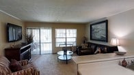 This single-floor residence offers a welcoming and functional layout centered around a cozy living area. The living room is carpeted and bathed in natural light, courtesy of a large window and a sliding glass door that opens to an outdoor balcony — ideal for enjoying fresh air and exterior views. The space features a textured ceiling and neutral-toned walls that create a warm, understated ambiance. Dark leather seating surrounds a glass-top coffee table, providing a comfortable and stylish arrangement for relaxing or entertaining. A large framed artwork adds a decorative focal point to one wall, enhancing the room's character. The sliding vertical blinds offer flexible light control and privacy. The balcony, visible through the glass door, provides additional outdoor living space. The overall design of the unit is practical and inviting, with a layout that maximizes the sense of openness on a single level. The combination of soft carpeting, abundant natural light, and thoughtful furnishings makes this a comfortable and well-appointed living space suited for everyday living.