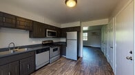 This single-floor unit offers a functional and well-appointed layout across several distinct living areas. The kitchen serves as a central highlight, featuring dark espresso-toned cabinetry with ample upper and lower storage, dark countertops, and a full suite of stainless steel appliances including a gas range, built-in microwave, dishwasher, and a full-size refrigerator. Wood-look vinyl plank flooring runs throughout the kitchen and living areas, lending a cohesive and easy-to-maintain aesthetic. The living area is bright and open, benefiting from a large multi-pane window that frames views of surrounding greenery and allows generous natural light to fill the space. A wall-mounted HVAC unit and baseboard heaters provide climate control throughout. The bedrooms are carpeted for comfort, each featuring windows with outdoor views and access to natural light; one bedroom includes a built-in closet with sliding doors. The bathroom is cleanly finished in white, featuring a single-sink vanity with cabinet storage, a mirror with overhead lighting, a toilet, and a full tub-shower combination with chrome fixtures. Overall, the unit presents a practical and comfortable living environment with a neutral color palette and consistent finishes throughout.