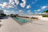 Located at 101 SW 1st St in Dania Beach, Florida, this rooftop amenity deck offers a refined, resort-inspired outdoor experience designed for both relaxation and social gathering. The centerpiece of the space is a generously sized swimming pool with crystal-clear turquoise water, finished with clean-edged porcelain tile decking in a neutral tone that reflects the South Florida sunshine. Shallow entry points and stainless steel handrails provide easy access throughout.

Surrounding the pool, an array of cushioned navy blue chaise lounges and circular lounge seating pods are thoughtfully arranged to maximize both comfort and open-sky exposure. Accent pieces in warm yellow and gold tones add pops of color against the predominantly white and grey palette. A dedicated spa zone with a shallow wading area and reclined loungers offers a more tranquil setting.

A wood-paneled pergola provides shaded retreat space, complemented by built-in planters filled with tropical foliage including bamboo, palms, and ornamental shrubs that frame the perimeter. Floor-to-ceiling glass walls with sliding doors connect the deck to an interior amenity space, bathed in natural light. The overall aesthetic is clean, contemporary, and unmistakably coastal.