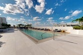 Located at 101 SW 1st St in Dania Beach, Florida, this rooftop amenity deck offers a refined, resort-inspired outdoor experience designed for both relaxation and social gathering. The centerpiece of the space is a generously sized swimming pool with crystal-clear turquoise water, finished with clean-edged porcelain tile decking in a neutral tone that reflects the South Florida sunshine. Shallow entry points and stainless steel handrails provide easy access throughout.

Surrounding the pool, an array of cushioned navy blue chaise lounges and circular lounge seating pods are thoughtfully arranged to maximize both comfort and open-sky exposure. Accent pieces in warm yellow and gold tones add pops of color against the predominantly white and grey palette. A dedicated spa zone with a shallow wading area and reclined loungers offers a more tranquil setting.

A wood-paneled pergola provides shaded retreat space, complemented by built-in planters filled with tropical foliage including bamboo, palms, and ornamental shrubs that frame the perimeter. Floor-to-ceiling glass walls with sliding doors connect the deck to an interior amenity space, bathed in natural light. The overall aesthetic is clean, contemporary, and unmistakably coastal.