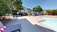 Located in Duluth, Georgia, this outdoor amenity space serves as a central gathering area for its residential community. The centerpiece is a generously sized swimming pool with wide concrete pool decking that provides ample room for lounging and socializing. Stainless steel entry rails offer safe access into the pool, which is complemented by a separate spa for relaxation.

The sundeck is well-appointed with rows of cushioned lounge chairs and shaded seating areas covered by large blue market umbrellas, ideal for enjoying sunny Georgia days. A pergola structure toward the rear of the space offers additional shaded gathering space. A ping-pong table adds a recreational element to the deck area.

Flanking one side of the pool is a classically styled building facade featuring decorative white columns, arched entryways, and stone accents set against a soft blue-gray exterior. Posted pool rules are prominently displayed near the entrance. The surrounding landscape is thoughtfully maintained with manicured shrubs, ornamental grasses, and mature shade trees that provide a sense of privacy and greenery. The entire space is enclosed by a fence for security, creating an inviting and functional outdoor retreat for residents.