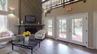 This single-floor space makes a strong architectural statement through its dramatic double-height ceilings, which lend an airy, expansive quality to the main living area. A prominent feature wall clad in diagonal wood planking serves as the focal point of the living room, anchoring a brick-hearth fireplace and a mounted flat-screen television above a substantial wooden mantel. The open layout connects the living area seamlessly to adjacent spaces, all unified by warm hardwood-style flooring throughout.

Large white-framed French doors line one wall of the living area, inviting generous natural light and providing views of the surrounding outdoor greenery. The elevated ceiling height is further emphasized by an interior upper-level balcony railing visible overhead, adding architectural depth to the space.

The kitchen features warm wood cabinetry extending to the ceiling, white appliances, and a peninsula counter with bar seating for casual dining. A separate seating area with soaring walls offers additional flexibility for layout and function.

Overall, the space balances rustic elements — exposed wood, brick, and natural textures — with clean, neutral tones, creating a warm yet open ambiance suitable for both relaxed everyday living and entertaining.