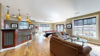 Located at 400 Blake St in New Haven, Connecticut, this single-floor community lounge space is thoughtfully designed for both relaxation and recreation. The open-plan layout is anchored by warm hardwood flooring that runs throughout, tying together the various functional zones seamlessly.

The entertainment area is a standout highlight, featuring a striking contemporary billiards table with a bold red felt surface and a sculptural curved base. Ample natural light pours in through large windows and glass doors that open to an outdoor patio area, enhancing the bright, welcoming atmosphere.

A comfortable lounge zone offers plush upholstered sofas and armchairs arranged around a glass coffee table, complemented by a wall-mounted flat-screen television — ideal for casual gatherings. Track lighting overhead and decorative pendant lights add warmth and dimension to the space.

The kitchenette area is well-appointed with rich wood cabinetry, dark speckled countertops, a stainless steel dishwasher, microwave, and a stylish tiled bar counter with bar stools illuminated by vibrant striped pendant lights. A small workstation area is also visible, providing a quiet nook for focused tasks.

The walls feature an earthy olive-green tone accented by framed artwork, contributing to a cohesive, contemporary aesthetic throughout this versatile and inviting community space.