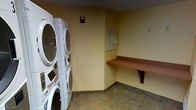 This single-floor laundry facility is designed for practical, efficient use. The room features stacked front-loading washers and dryers along one wall, maximizing the use of vertical space while keeping the floor area open and easy to navigate. The walls are painted in a warm yellow tone, complemented by a slate-style tile floor that adds durability and ease of maintenance. A wall-mounted wooden folding shelf provides a convenient surface for sorting or staging laundry. Informational postings are displayed on the wall, likely outlining usage guidelines or instructions for the machines. An electrical outlet is also visible on the adjacent wall, offering additional utility. The space is straightforward in layout, with no excess furnishings, keeping the focus on functionality. Overall, the laundry room presents a clean, no-frills environment well-suited to serving the everyday needs of residents or guests.