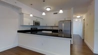 This single-floor residence offers a well-organized open-concept layout, as revealed by the floor plan's clear delineation of living, kitchen, and private spaces. Rich hardwood flooring flows consistently throughout the main living areas, creating a warm and cohesive aesthetic.

The kitchen serves as a central highlight of the space, designed with a large peninsula that provides both additional workspace and a natural division between the kitchen and adjoining living areas. White shaker-style cabinetry lines the walls from counter to ceiling, offering generous storage capacity. Dark granite countertops provide a striking contrast against the bright cabinetry and white walls. Stainless steel appliances, including a French door refrigerator, electric range, and built-in microwave, complete the modern kitchen ensemble. A trio of pendant lights hang above the peninsula, supplemented by flush-mount ceiling fixtures that ensure the space is well-lit.

The floor plan indicates additional rooms branching off from the main living area, including what appear to be bedrooms and a bathroom, each accessible via a central corridor. The overall ambiance of the space is clean, bright, and contemporary.