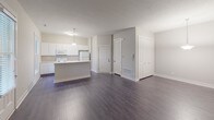 This single-floor unit is designed with a clean, functional layout that emphasizes an open and connected living environment. The kitchen serves as a central focal point, featuring crisp white cabinetry extending to the upper walls for ample storage, white appliances including a refrigerator, electric range, and over-the-range microwave, and light countertops throughout. A peninsula island with a built-in sink anchors the kitchen, providing additional workspace and a natural gathering point. A pendant light hangs above the island, complementing the natural light that enters through a nearby window. The kitchen flows seamlessly into the open living area, which is finished with dark hardwood-style flooring that extends throughout the main space, creating a cohesive and polished look. The living area is generously sized with neutral-toned walls and a textured ceiling, offering a versatile canvas for various arrangements. A full bathroom features a white vanity, toilet, and a tub-shower combination with a curved curtain rod. Closet space is accessible from the main living area, and the entry is clearly defined, contributing to the unit's organized flow.