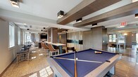 This single-floor amenity lounge is designed with a contemporary aesthetic, combining rich wood tones, textured wall finishes, and exposed beam ceiling details to create a refined yet inviting atmosphere. The space is organized into several distinct functional zones that flow seamlessly into one another.

At the center of the room, a full-size pool table with blue felt serves as a focal point, positioned beneath a striking ceiling treatment of parallel dark wood beams fitted with square black pendant fixtures. Adjacent to the billiards area, a comfortable lounge zone features neutral-toned sofas, armchairs, and low side tables arranged on a textured area rug, with a wall-mounted flat-screen television nearby.

A wet bar area is equipped with stainless steel under-counter refrigeration units, a sink, white countertops, and a decorative tile backsplash, providing a functional serving space ideal for gatherings. High-top communal tables with bar-height seating offer additional space for dining or socializing.

Natural light enters through several large windows, brightening the lounge and billiards areas. Wood-look tile flooring runs throughout, complementing the warm cabinetry and wall paneling. The overall ambiance is polished and social, well-suited for entertainment and community interaction.
