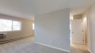 This single-floor residential space presents a clean, understated aesthetic defined by neutral wall tones, white trim, and smooth ceilings throughout. The main living area is generously proportioned, offering an open and airy feel that invites flexible furniture arrangements. Plush wall-to-wall carpeting in a light gray hue runs throughout, adding warmth and comfort underfoot.

Natural light is a key feature of this space, with a wide set of multi-pane windows along one wall framing views of lush greenery outside. These windows not only brighten the interior but also create a pleasant visual connection to the outdoors. A wall-mounted air conditioning unit sits beneath the windows, complemented by baseboard heating that runs along the perimeter — together providing year-round climate comfort.

A partial interior wall divides the space, creating a natural sense of separation between living areas while maintaining an open flow. Through an adjacent doorway, additional rooms are accessible, suggesting a well-organized and functional layout. The overall color palette — warm beige, soft gray, and crisp white — lends the space a calm, neutral ambiance that provides an ideal blank canvas for personalization.