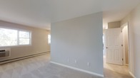 This single-floor residential space presents a clean, understated aesthetic defined by neutral wall tones, white trim, and smooth ceilings throughout. The main living area is generously proportioned, offering an open and airy feel that invites flexible furniture arrangements. Plush wall-to-wall carpeting in a light gray hue runs throughout, adding warmth and comfort underfoot.

Natural light is a key feature of this space, with a wide set of multi-pane windows along one wall framing views of lush greenery outside. These windows not only brighten the interior but also create a pleasant visual connection to the outdoors. A wall-mounted air conditioning unit sits beneath the windows, complemented by baseboard heating that runs along the perimeter — together providing year-round climate comfort.

A partial interior wall divides the space, creating a natural sense of separation between living areas while maintaining an open flow. Through an adjacent doorway, additional rooms are accessible, suggesting a well-organized and functional layout. The overall color palette — warm beige, soft gray, and crisp white — lends the space a calm, neutral ambiance that provides an ideal blank canvas for personalization.