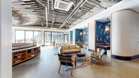 This clubhouse in Baltimore, MD offers a generously proportioned single-floor gathering space defined by a sophisticated blend of industrial and contemporary design elements. The exposed ceiling reveals structural framework alongside ductwork and HVAC systems, while wood-slat ceiling installations in select zones add warmth and acoustic texture. Polished concrete floors extend throughout, unifying the open layout.

The main lounge area features multiple seating arrangements anchored by patterned area rugs, mid-century-inspired wood-frame furniture, and a distinctive curved built-in bookcase with a marble top, creating a central social hub. A bold blue accent wall adorned with a graphic koi-and-diver mural serves as a focal point, framed by warm pendant lighting. Additional lounge zones provide intimate gathering spaces with white sofas and tropical potted plants.

A dedicated conference room features a large oval table, upholstered seating, a display screen, and a geometric-patterned wall treatment with an overhead statement pendant. Floor-to-ceiling sliding glass doors on the waterfront-facing wall flood the space with natural light and open to an outdoor terrace offering direct views of Baltimore's harbor. The entry area includes a reception desk and curated artwork, setting a polished, design-forward tone throughout.