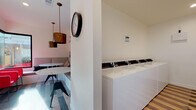 Located at 1500 Alamo Dr in Vacaville, California, this single-floor amenity space is thoughtfully designed to combine functionality with contemporary style. The space is divided into two distinct areas: a vibrant lounge and a fully equipped laundry facility.

The lounge area makes a strong design statement, featuring striking marble-top tables paired with built-in upholstered banquette seating and bold red accent chairs. An eye-catching abstract watercolor mural anchors one wall, while copper-toned pendant lights hang overhead, adding warmth and character. Large black-framed windows flood the space with natural light and provide views of the surrounding outdoor courtyard. The multi-toned striped wood-look flooring runs throughout, tying the areas together cohesively.

Adjacent to the lounge, the laundry facility is clean and efficiently organized, with rows of commercial-grade washers and dryers lining both walls. A fold-down countertop provides a convenient surface for sorting and folding laundry. A framed watercolor artwork adorns the wall, maintaining the aesthetic continuity of the overall space. Recessed lighting and neutral white walls keep the laundry area bright and practical, making it a well-considered and inviting shared resource.