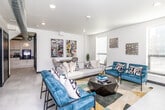 This single-floor space showcases a contemporary industrial aesthetic, blending clean modern finishes with distinctive exposed silver ductwork that runs throughout the ceiling. The main lounge area is generously proportioned, featuring large windows with slatted blinds that allow ample natural light to filter in, creating a bright and welcoming atmosphere. Light-toned large-format tile flooring extends seamlessly throughout, contributing to the open, airy feel of the space.

The lounge is furnished with a neutral linen sofa, paired with striking teal upholstered accent chairs featuring bold geometric-patterned cushions, all anchored by a dark coffee table and a patterned area rug with yellow and grey tones. Curated wall art and decorative accents add personality throughout. A wall-mounted flat-screen television is positioned opposite the seating area, supported by a gold-toned media console.

A dedicated coffee bar area is thoughtfully equipped with white cabinetry, dark countertops, a utility sink, and a professional-grade coffee machine, accented by a glowing neon "Coffee" sign that adds a playful touch. Additional rooms with dark-framed doorways branch off from the main area, alongside an accessible restroom, offering functional versatility within the space.