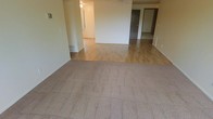This single-floor space offers a clean, functional layout with a welcoming open-plan living area at its core. The main living zone features neutral-toned carpet that transitions seamlessly into hardwood-style flooring, delineating distinct areas within the open floor plan. The walls are finished in a light, neutral palette throughout, contributing to a bright and airy atmosphere.

Ceiling fans are installed in the main living areas, providing both ventilation and a casual aesthetic. Floor vents are visible along the baseboards, indicating a central HVAC system for year-round climate control. The open layout allows for flexible furniture arrangement and a natural flow between spaces.

A partial wall or breakfast bar feature separates the main living area from an adjacent space, adding architectural interest while maintaining an open feel. Beyond the living area, a hallway leads to additional rooms, suggesting a private sleeping or utility wing. Doorways visible in the background hint at multiple rooms, offering practical separation between living and private spaces.

Overall, this space presents a simple, move-in-ready environment with a neutral design foundation, making it highly adaptable to a variety of interior styles and uses.