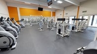 This fitness facility is a single-floor gym designed to deliver a complete workout experience in a clean, open environment. The main workout floor is expansive, featuring durable rubber flooring in a neutral gray tone that provides both safety and comfort underfoot. The space is outfitted with a wide array of white-framed strength training machines, including cable systems, plate-loaded machines, and adjustable benches, all thoughtfully arranged to allow for easy navigation and safe use. Cardio equipment, including treadmills, lines the perimeter of the space.

The interior design strikes a bold yet functional aesthetic, with vibrant orange accent walls that energize the atmosphere and contrast sharply with the bright white walls and ceiling. Overhead, large fluorescent light panels and recessed lighting ensure the entire floor is evenly and brightly illuminated. Mounted televisions are suspended from the ceiling, offering entertainment throughout workouts.

Floor-to-ceiling glass panels and windows along one side of the facility allow natural light to pour into the space, contributing to an open and inviting ambiance. The high ceilings further enhance the sense of openness and airiness, making this gym both practical and motivating for users of all fitness levels.
